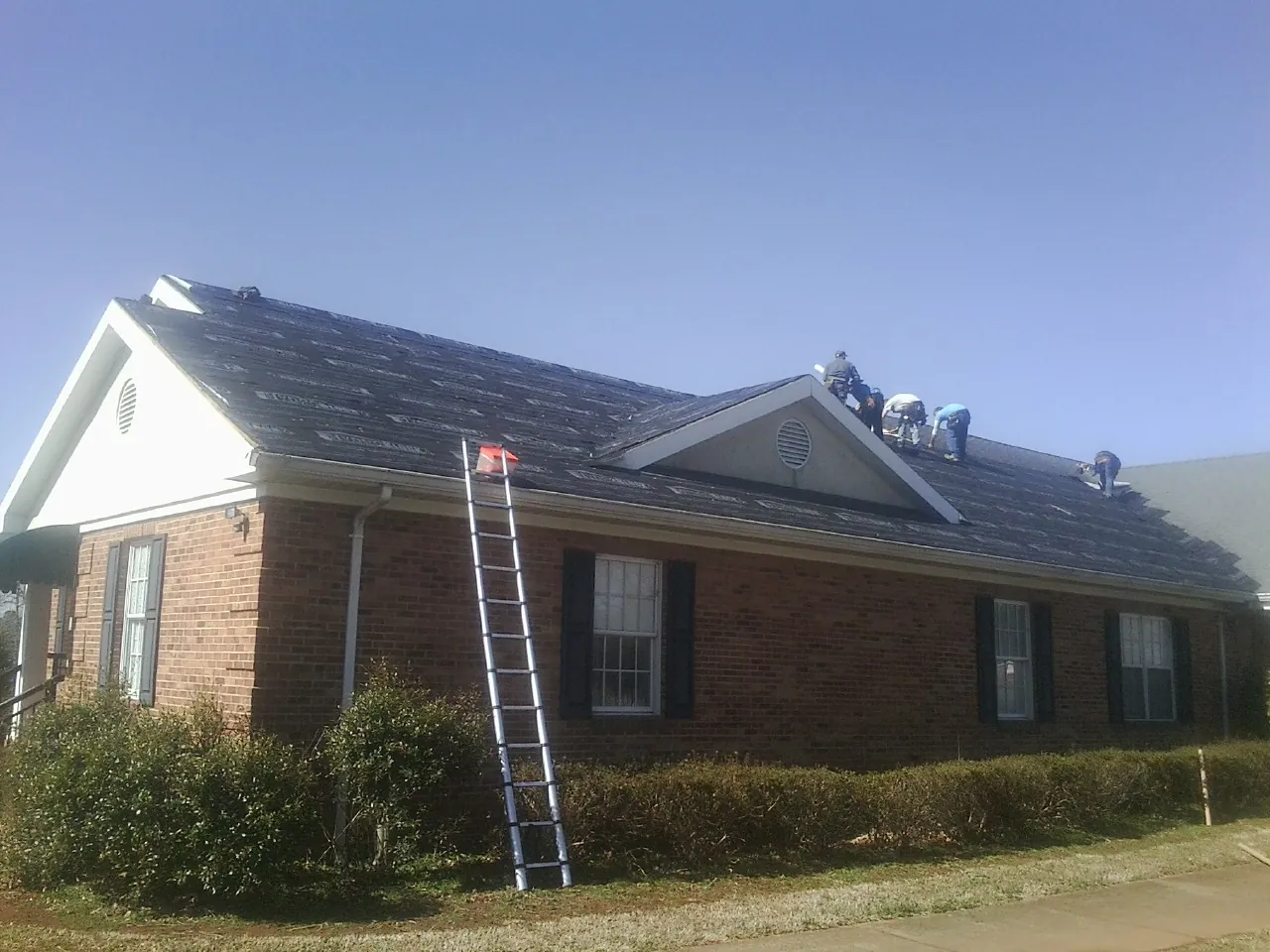 Crew tearing shingles off a brick ranch roof with a ladder set — Metal Roof Repair work by our crew in Manning